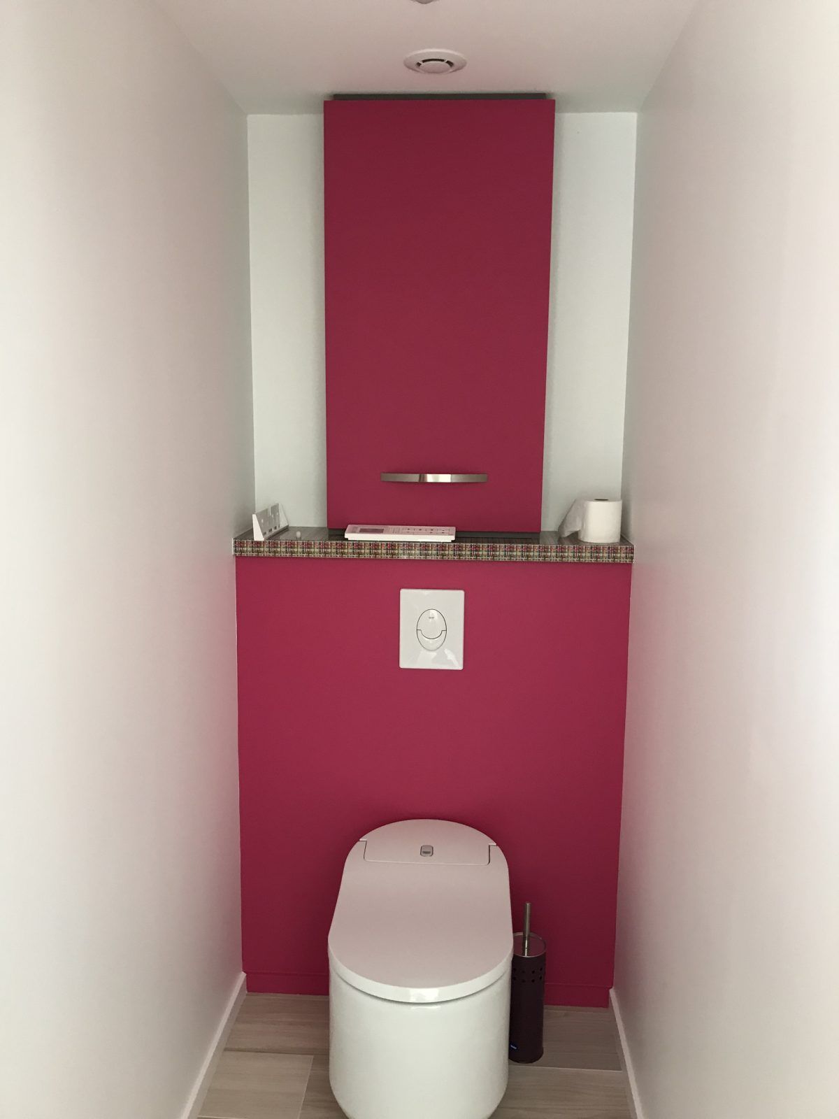Une petite salle de toilettes avec des toilettes blanches adossées à un mur magenta éclatant, une étagère en granit et un meuble assorti.