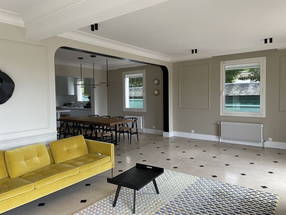 Un salon lumineux comprend un canapé jaune, un tapis à motifs, une table basse noire et un coin repas ouvert attenant.