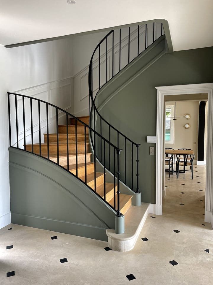 Un escalier courbe avec des marches en bois, une rampe en fer noir et des murs de soubassement vert olive dans une maison élégante aux tons neutres.