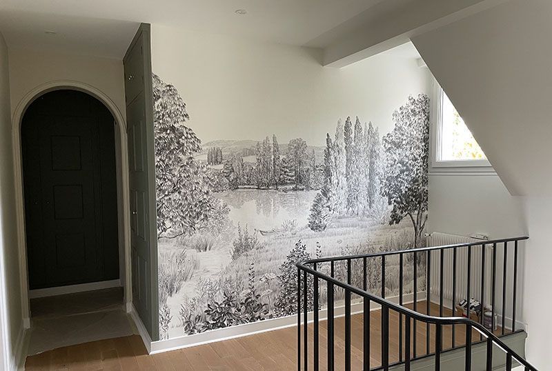Un palier d'escalier avec parquet, rampes noires et une grande fresque monochrome représentant un paysage au mur.