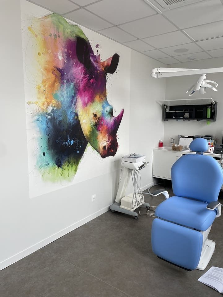 Une fresque murale à l'aquarelle aux couleurs vives représentant une tête de rhinocéros sur un mur blanc dans un cabinet dentaire, avec un fauteuil de patient bleu.