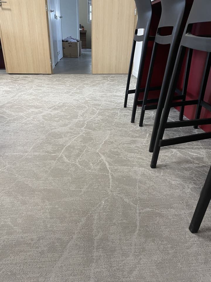 Gros plan sur un tapis gris à motifs au sol, à côté des pieds de plusieurs tabourets de bar foncés, dans un bureau ou une cuisine.