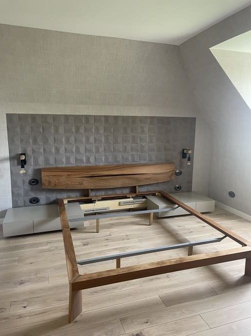 Un cadre de lit en bois est placé devant un mur carrelé gris, avec une tête de lit en bois suspendue et deux tables de chevet.