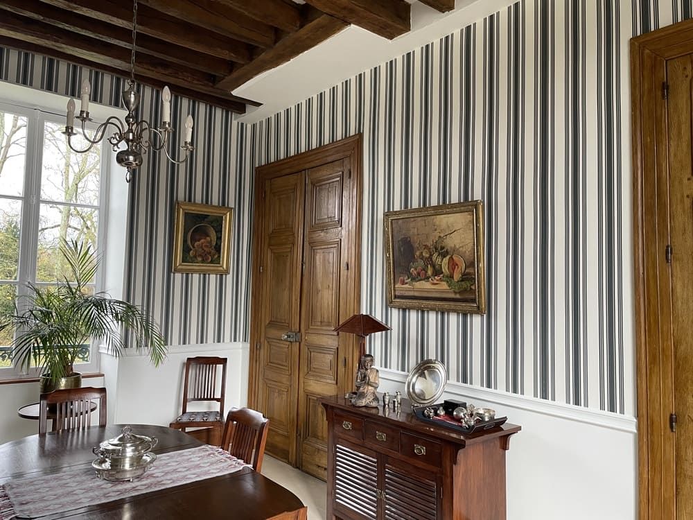 Salle à manger avec papier peint à rayures, portes à panneaux de bois, lustre et buffet en bois présentant des objets décoratifs.