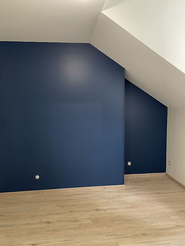 Une pièce vide avec un sol clair imitant le bois et des murs bleu foncé sous un plafond mansardé.