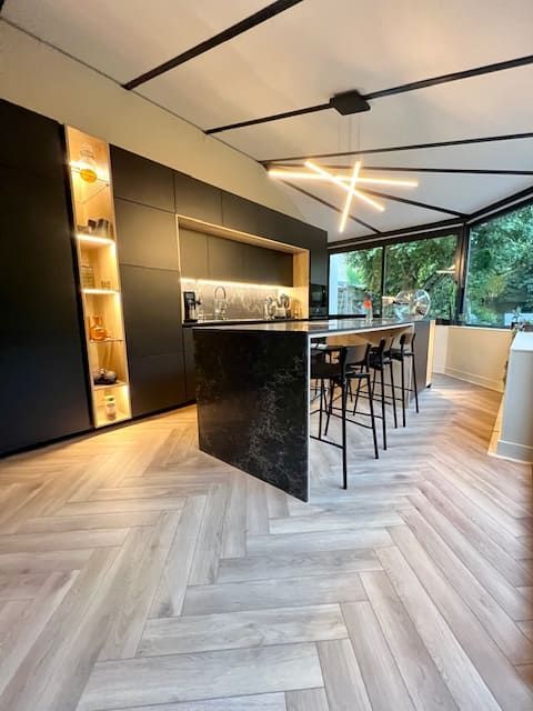 Cuisine moderne avec des armoires foncées mates, un îlot central en marbre avec tabourets de bar et un parquet clair à chevrons.