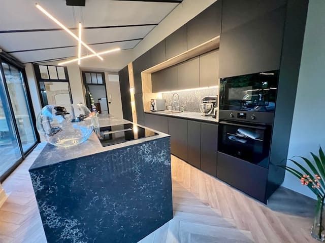 Cuisine moderne avec des armoires aux tons foncés mats, un îlot central en pierre, un parquet clair et un éclairage linéaire minimaliste.