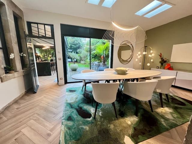 Salle à manger moderne avec une table ovale, des chaises blanches et un tapis à motifs verts audacieux, donnant sur un jardin-terrasse.
