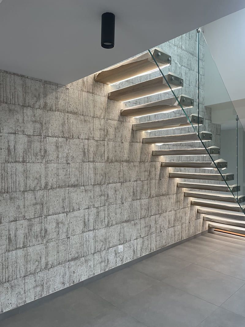 Escalier flottant en bois avec éclairage sous les marches, adossé à un mur en pierre texturée, à côté d'une balustrade en verre.