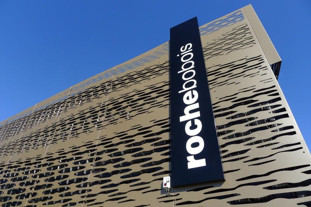 Une enseigne verticale noire avec le logo blanc « rochebobois » montée sur un bâtiment texturé de couleur claire sous un ciel bleu.