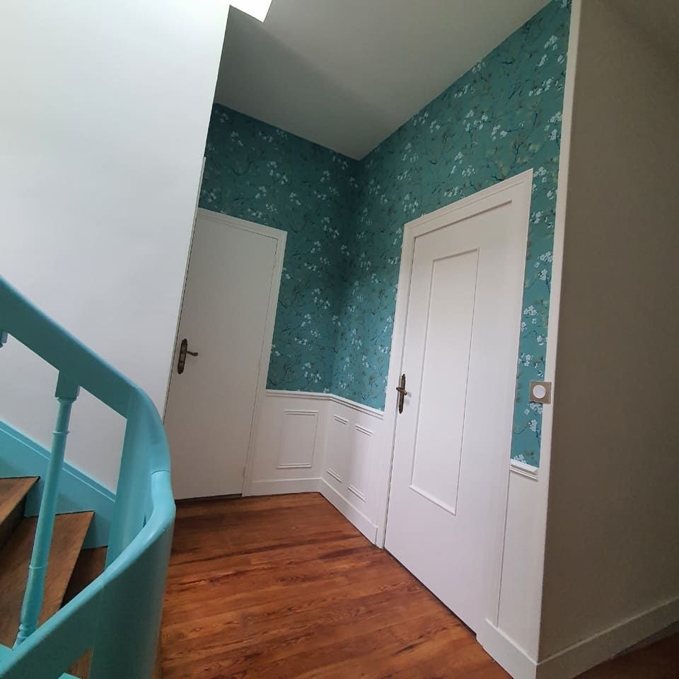 Un couloir avec parquet, papier peint fleuri turquoise au-dessus de lambris blancs, deux portes blanches et une rampe turquoise vif.