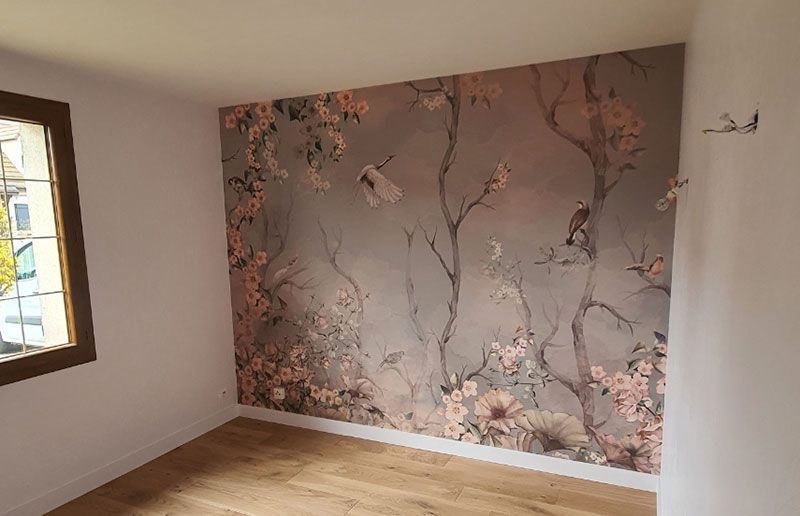 Une pièce avec un mur orné d'une fresque murale représentant un arbre et des fleurs dans des tons rose poudré, gris et blanc, un sol clair et une fenêtre.