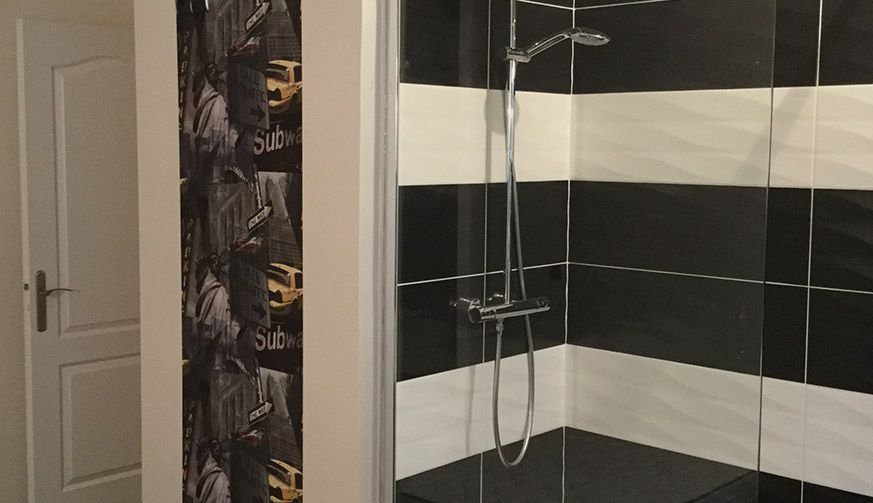Une douche de salle de bain avec des murs carrelés horizontalement en noir et blanc, jouxtant un mur recouvert de papier peint à motifs et une porte fermée.