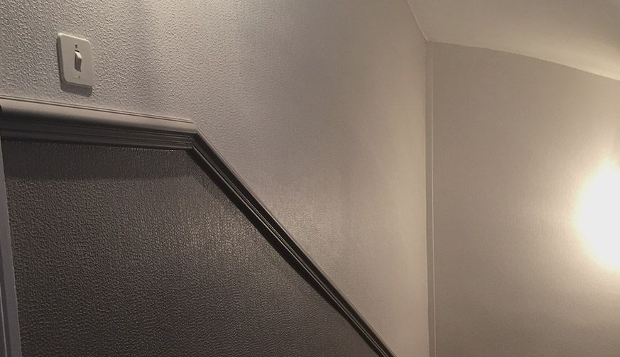 Un mur blanc avec un interrupteur et une rampe d'escalier diagonale de couleur sombre.
