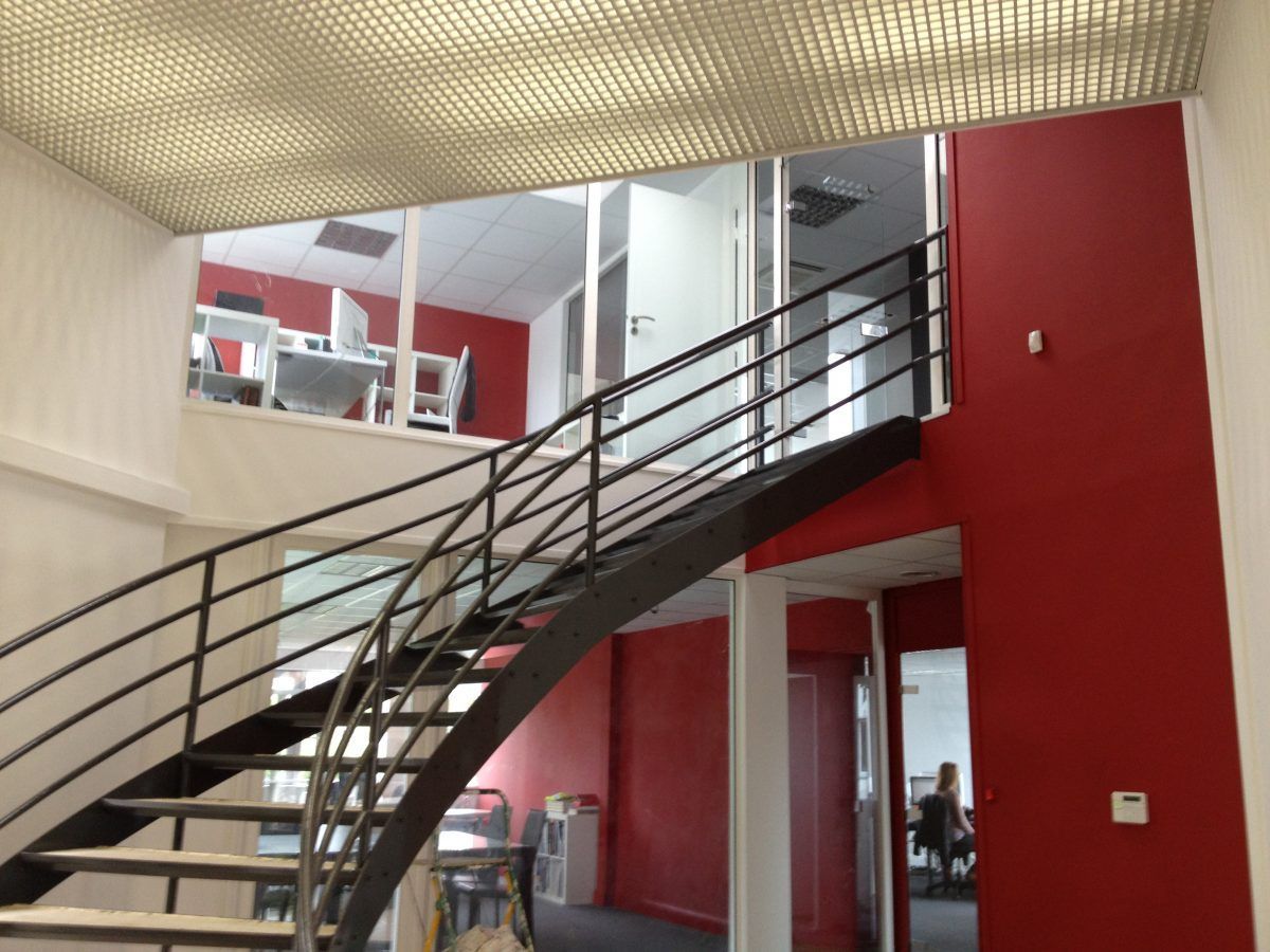 Un escalier métallique incurvé mène à un étage de bureaux supérieur aux murs rouges et au plafond à motif quadrillé.