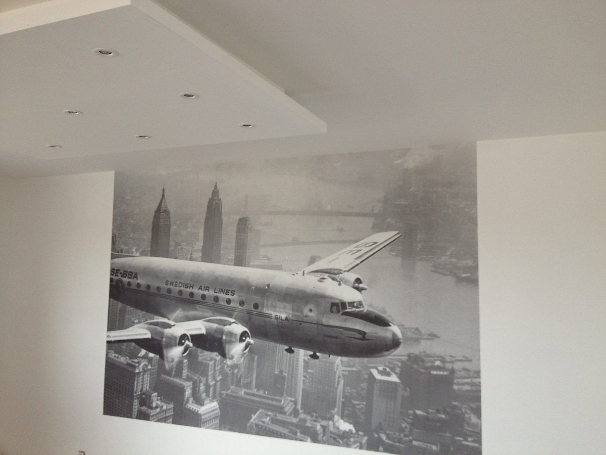 Une fresque murale en noir et blanc représentant un avion de ligne vintage survolant l'horizon d'une ville du milieu du XXe siècle.