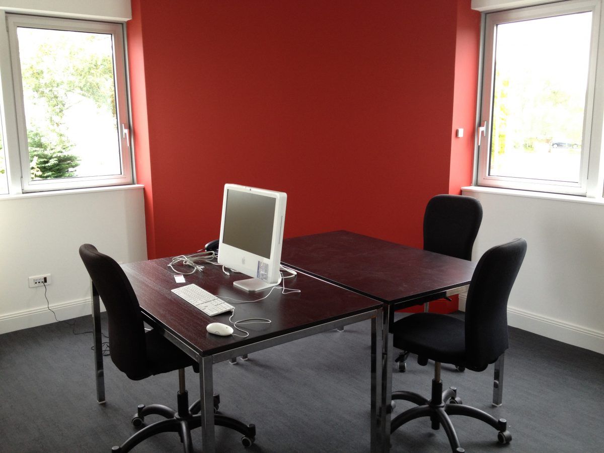 Un poste de travail informatique avec deux chaises de bureau noires autour d'un bureau, adossé à un mur rouge vif entre deux fenêtres.