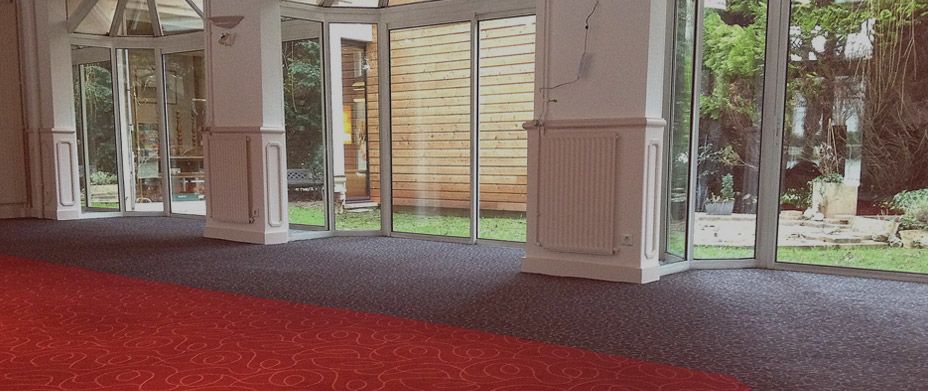 Un tapis rouge traverse une pièce moderne et lumineuse, dotée de grandes fenêtres donnant sur un jardin et un mur extérieur en bois.