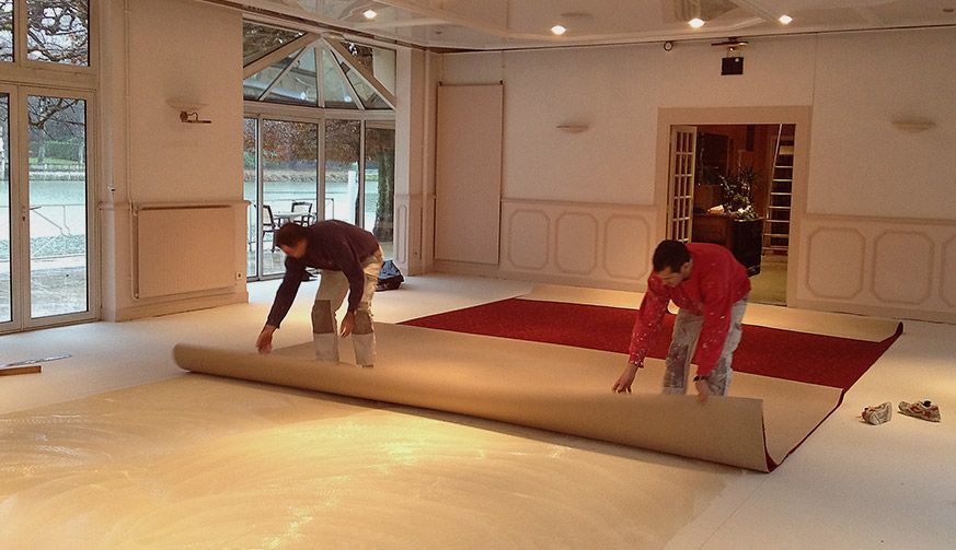 Deux ouvriers déroulent un long tapis beige sur un sol blanc dans une grande pièce lumineuse.