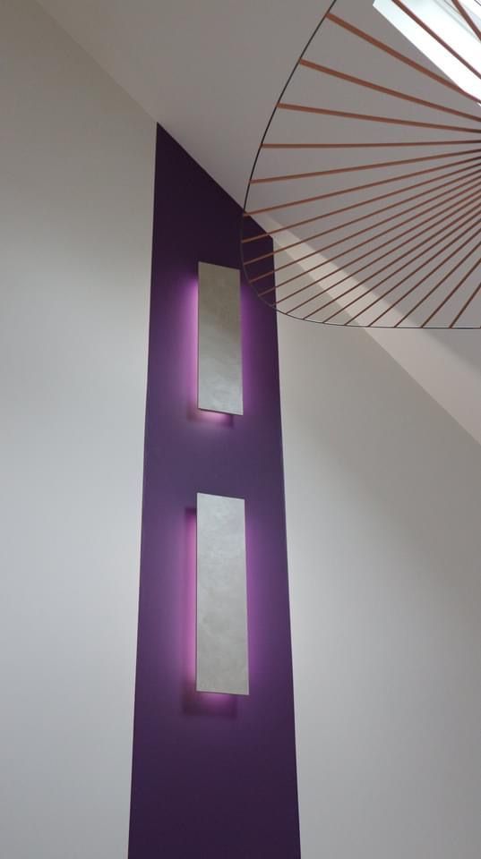 Deux appliques murales rectangulaires fixées sur une bande verticale violette sur un mur blanc, diffusant une lumière douce et chaude.