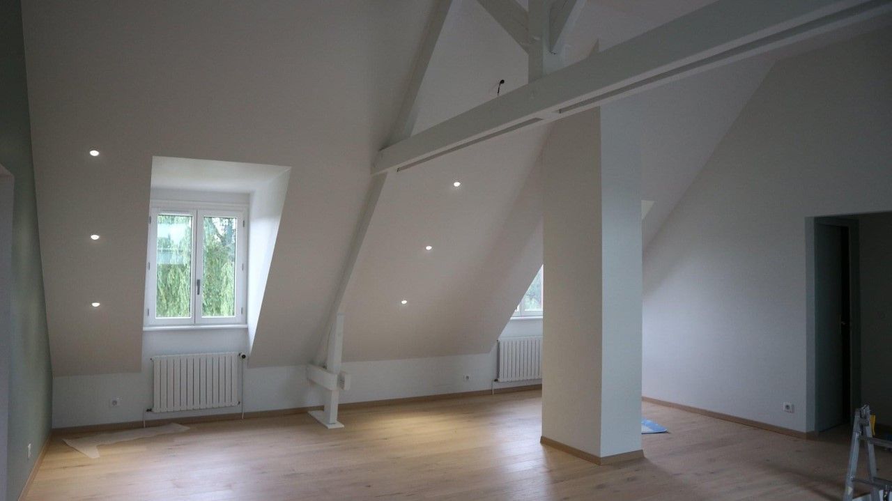 Une pièce mansardée lumineuse et vide, avec parquet, murs blancs, plafonds mansardés, poutres apparentes et spots encastrés.