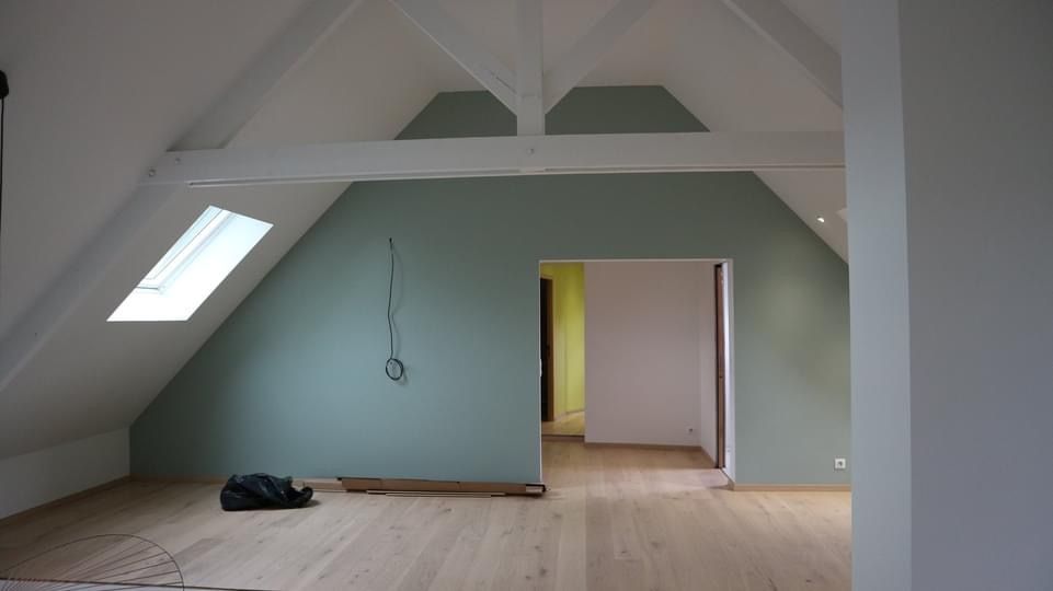 Une pièce mansardée avec parquet clair, murs vert sauge, plafonds blancs en pente, poutres apparentes et puits de lumière.
