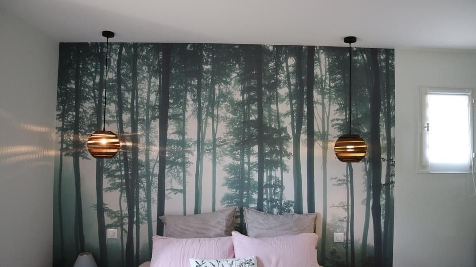 Un mur de la chambre est recouvert d'un papier peint à motif de forêt, avec deux suspensions lumineuses au-dessus d'un lit agrémenté d'oreillers roses et gris.