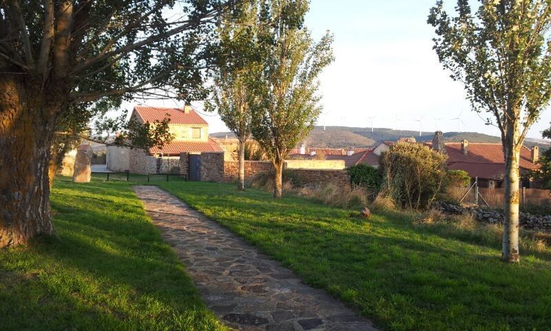 Un sendero de piedra serpentea a través de un parque cubierto de hierba hacia un pueblo con casas de ladrillo y árboles.