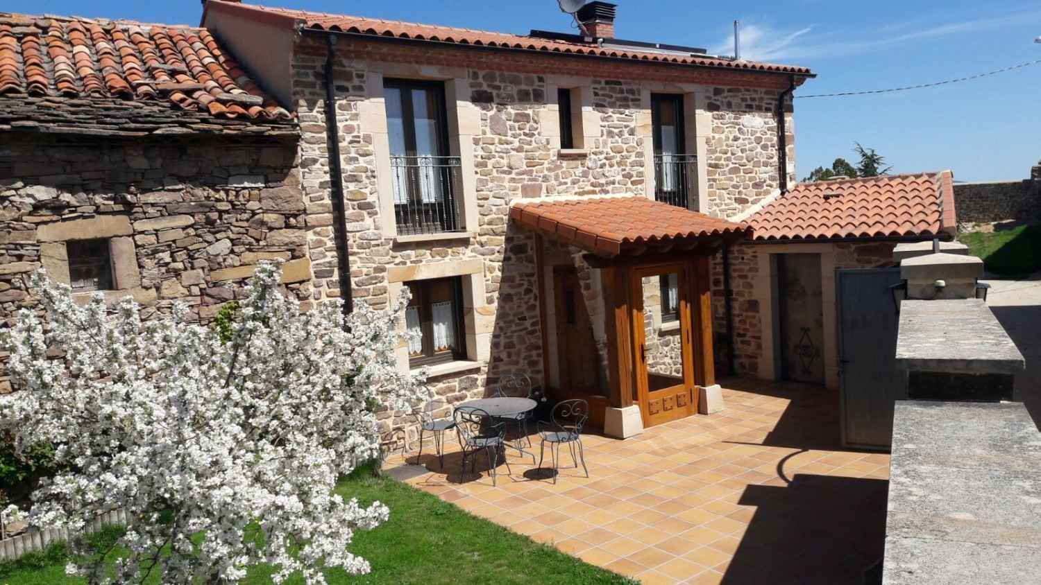 Una casa de piedra con tejado de tejas, patio y un árbol en flor delante, bajo un cielo azul despejado.
