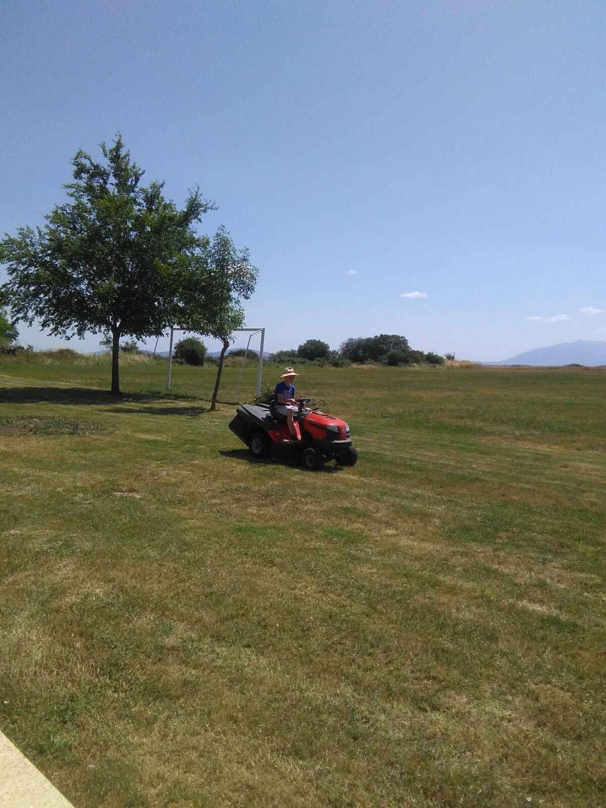 Una persona conduce una cortadora de césped roja a través de un amplio campo cubierto de hierba bajo un cielo azul.