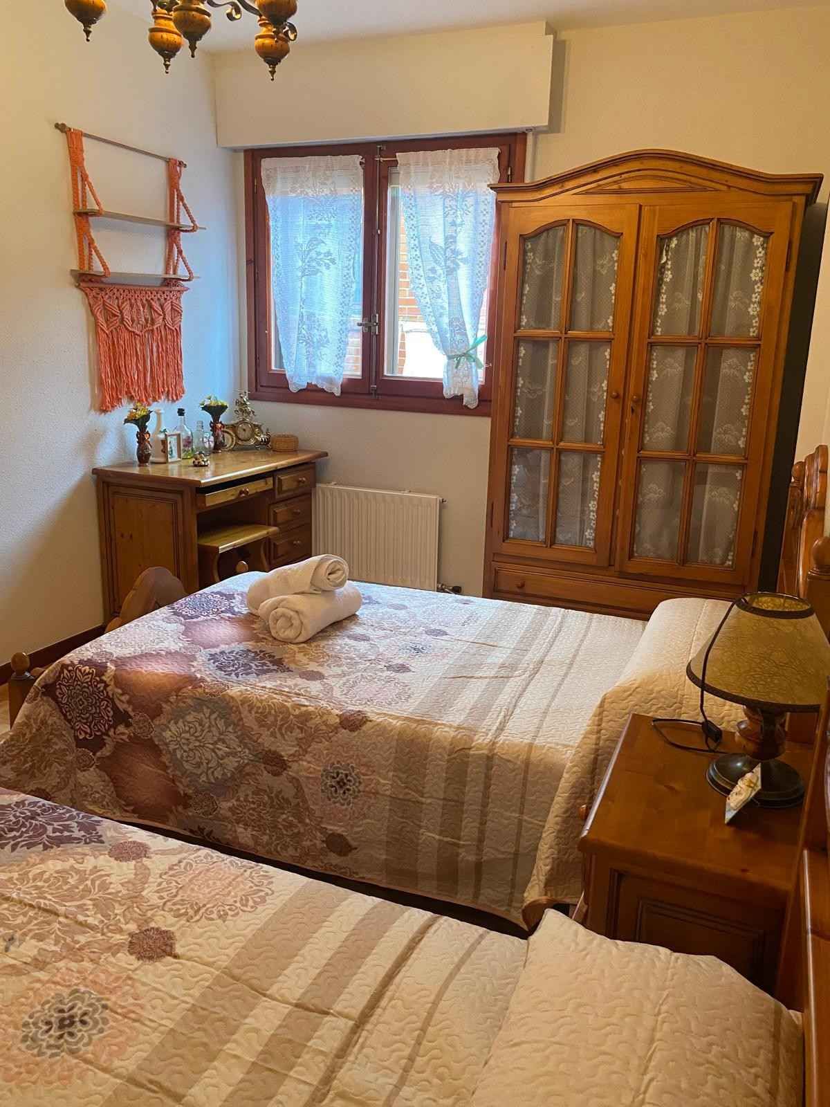 Un dormitorio con dos camas, un armario de madera, un escritorio con silla y cortinas transparentes en la ventana.