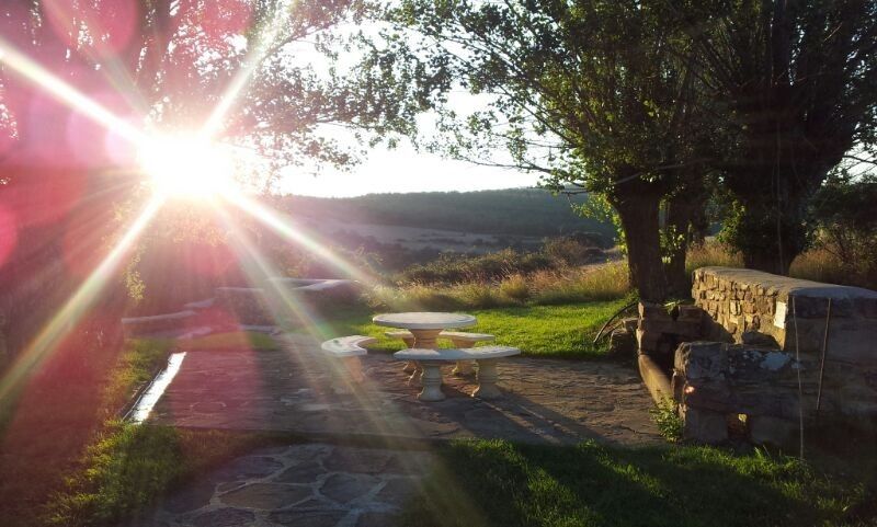 Un patio de piedra con mesa y bancos al atardecer, rodeado de árboles y un paisaje rural.