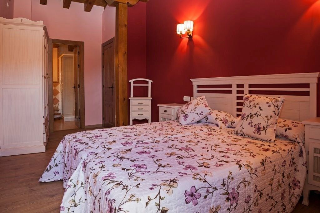 Un dormitorio con una cama con una colcha floral contra una pared roja, muebles de madera blanca y vigas de techo a la vista.