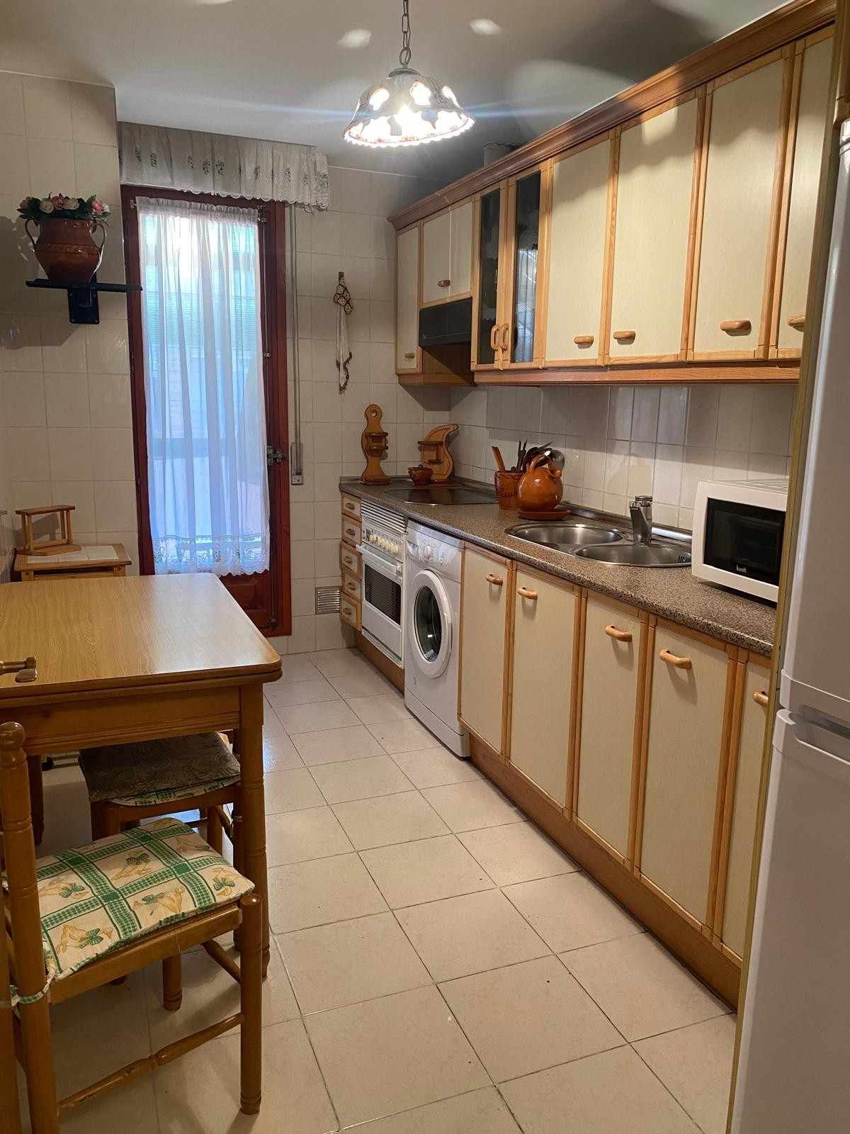 Una cocina alargada con armarios de tonos claros, lavadora, fregadero y mesa de madera con sillas.