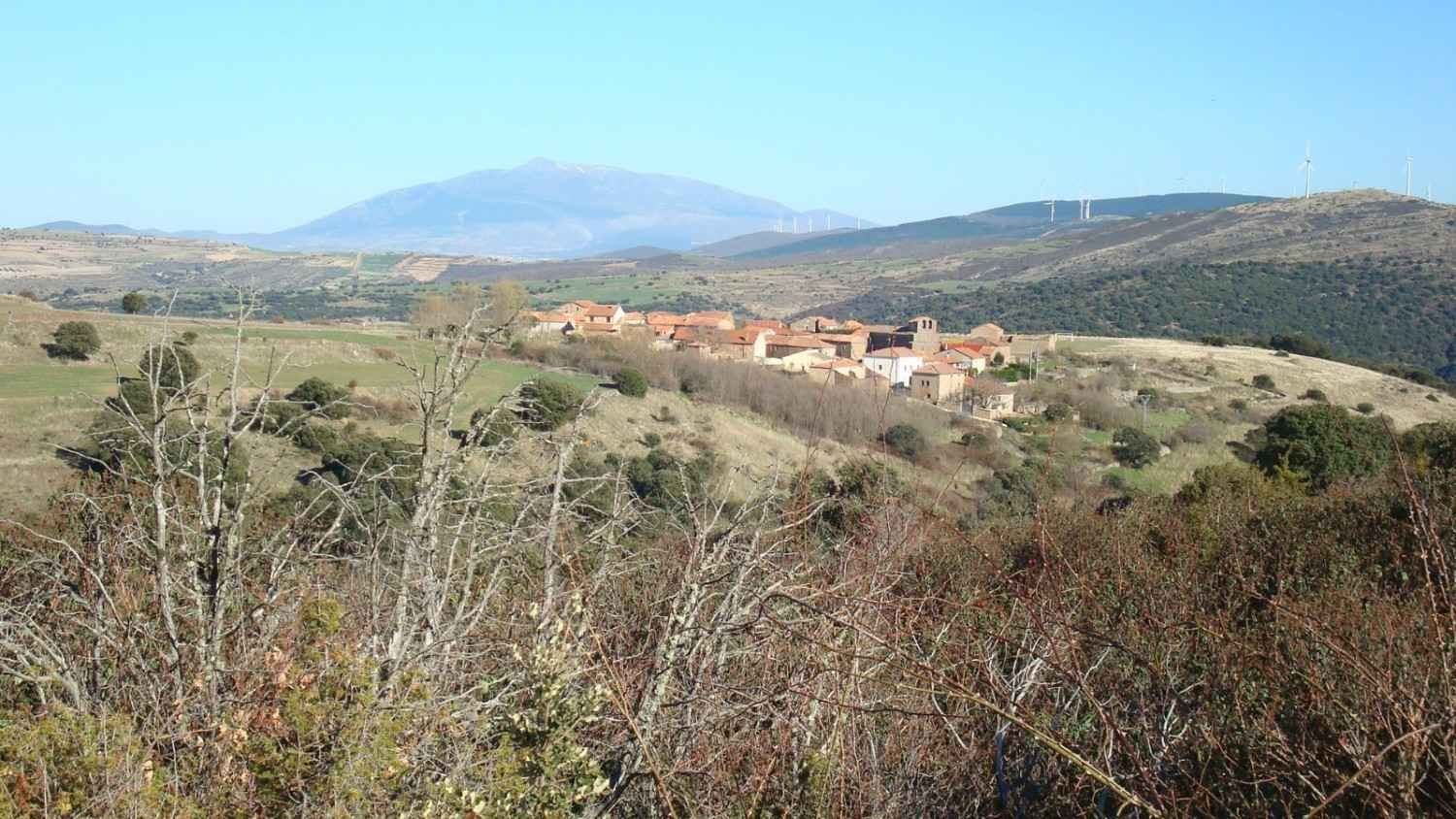 Un pequeño pueblo con edificios de tejados de terracota enclavado en un paisaje montañoso bajo un cielo azul despejado.