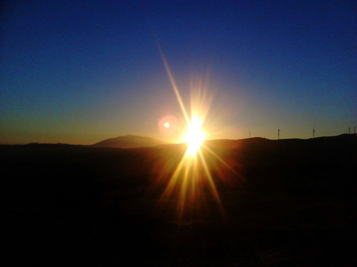 Un sol brillante se eleva sobre una oscura cordillera recortada contra un cielo de un azul intenso degradado.