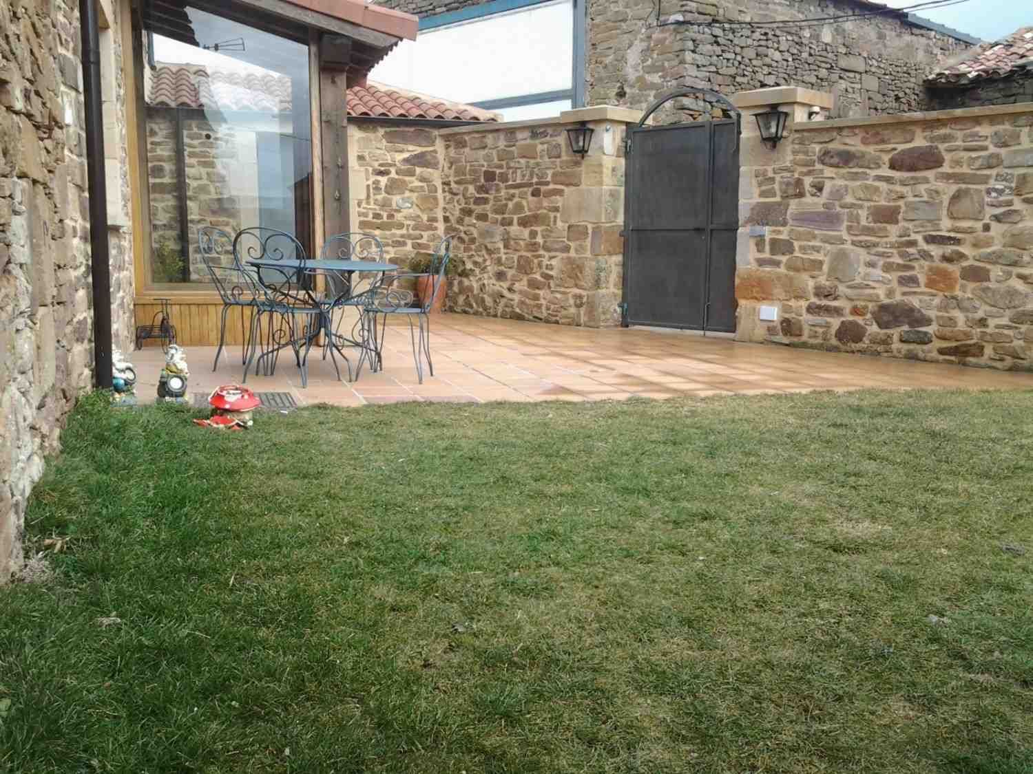 Un patio de piedra con una mesa y sillas de bistró se encuentra junto a un edificio con paredes de cristal y un muro.