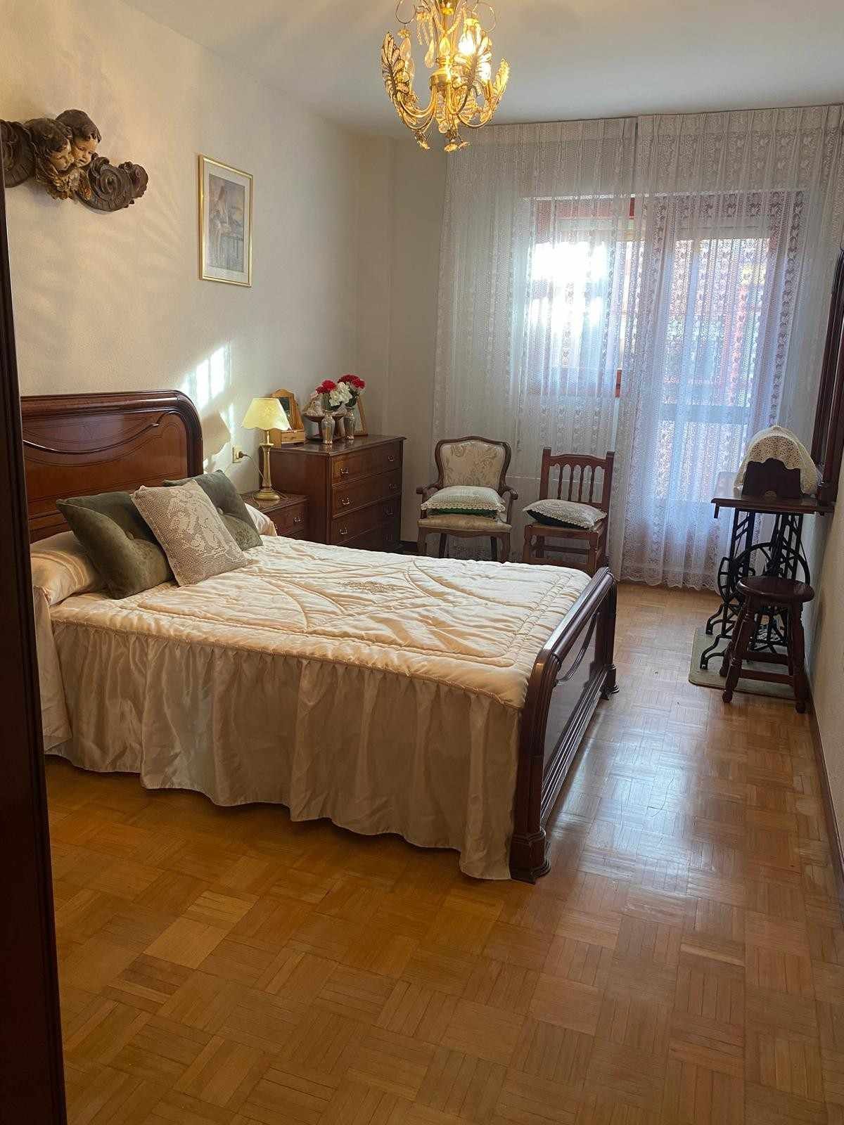 Un dormitorio con una cama de madera, una colcha blanca con volantes, una pequeña cómoda de madera.