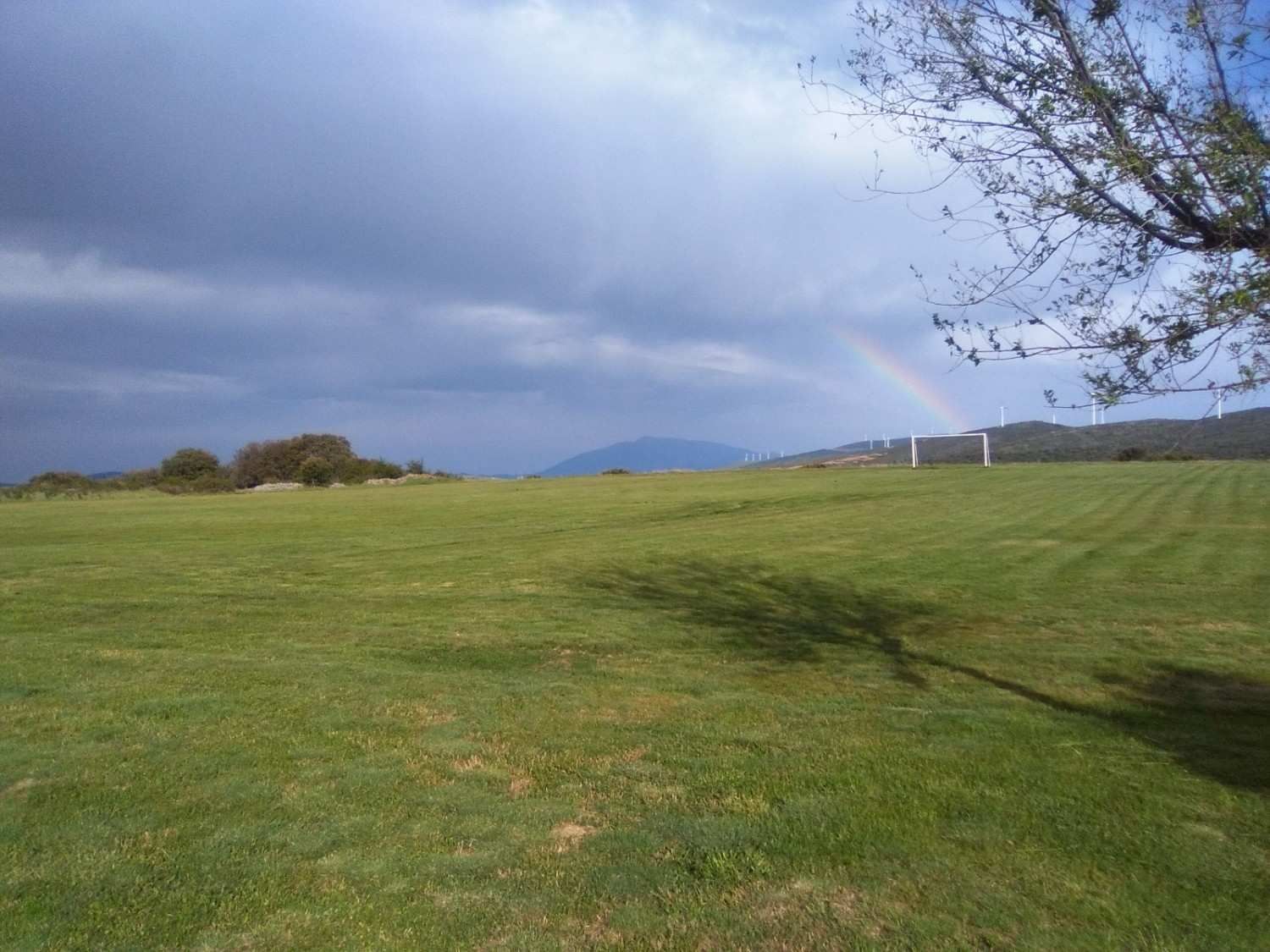 Un extenso campo cubierto de hierba bajo un cielo nublado, con un tenue arcoíris visible a lo lejos cerca de una colina.