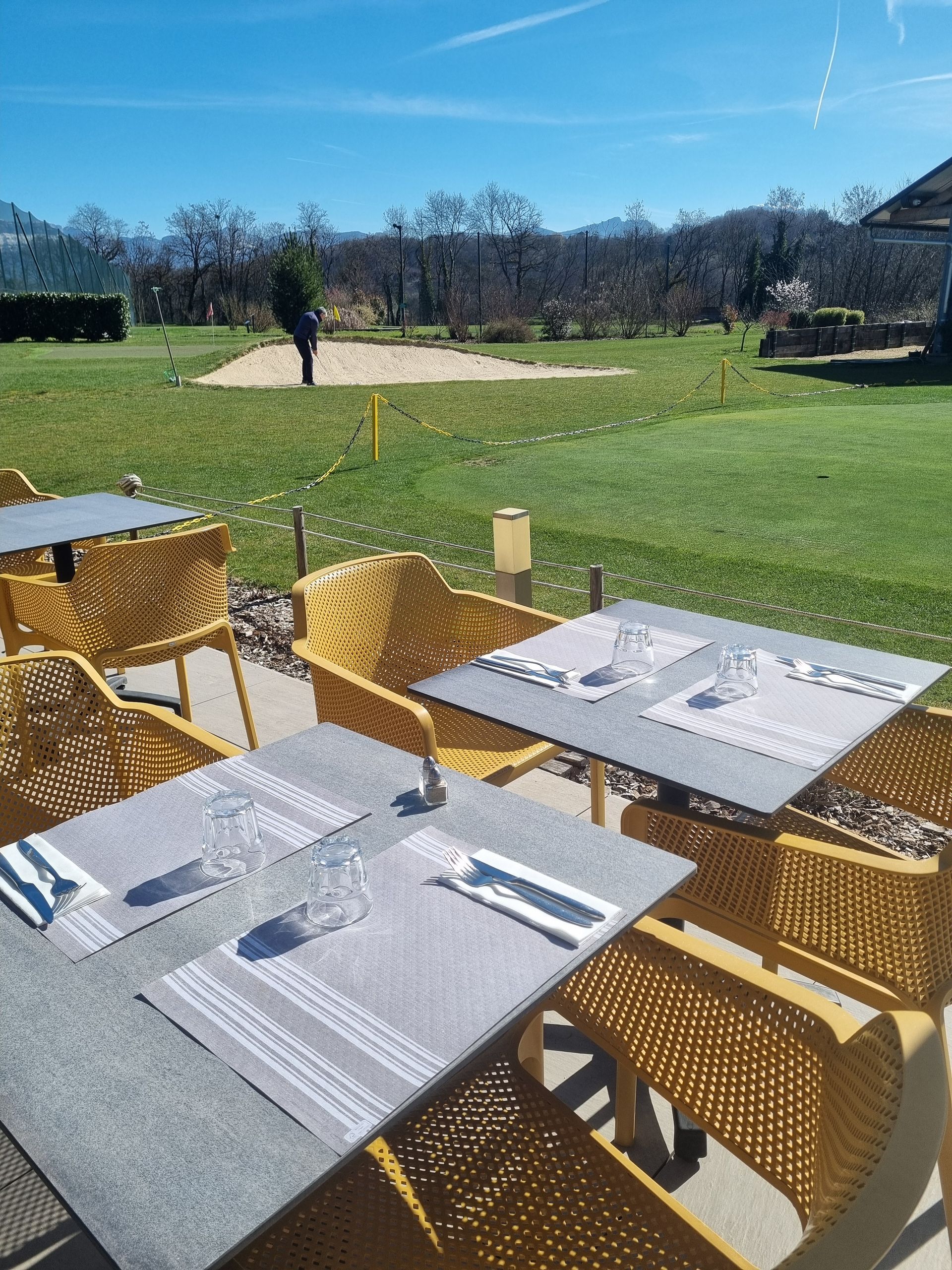 Tables de café en plein air et chaises en osier sur une terrasse ensoleillée donnant sur un champ verdoyant et des arbres.