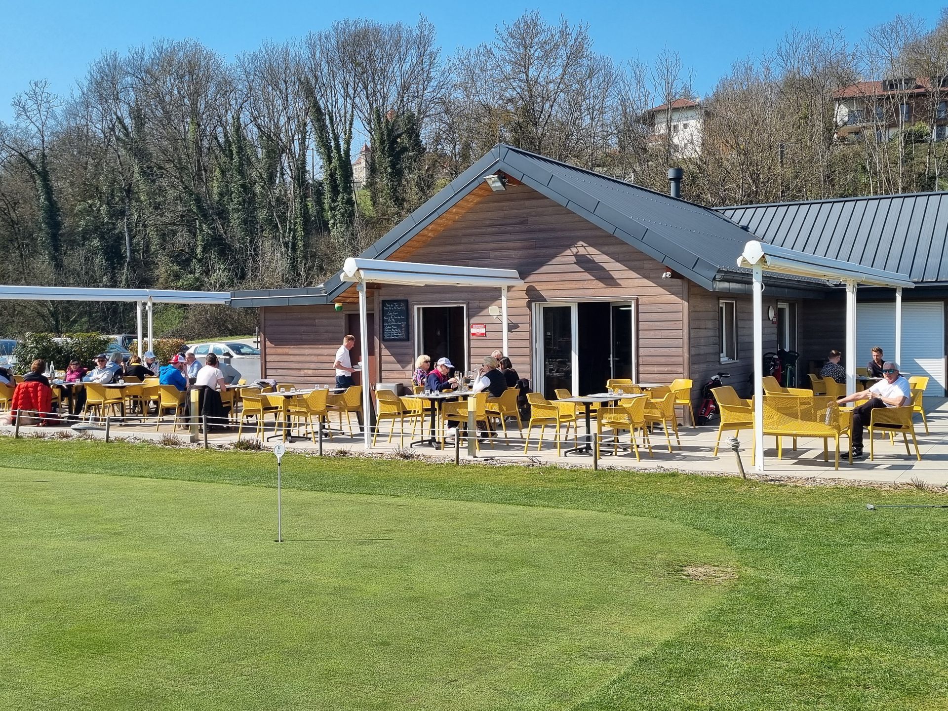 Terrasse extérieure de café jouxtant un petit bâtiment en bois, avec des tables et des chaises jaunes par une journée ensoleillée.