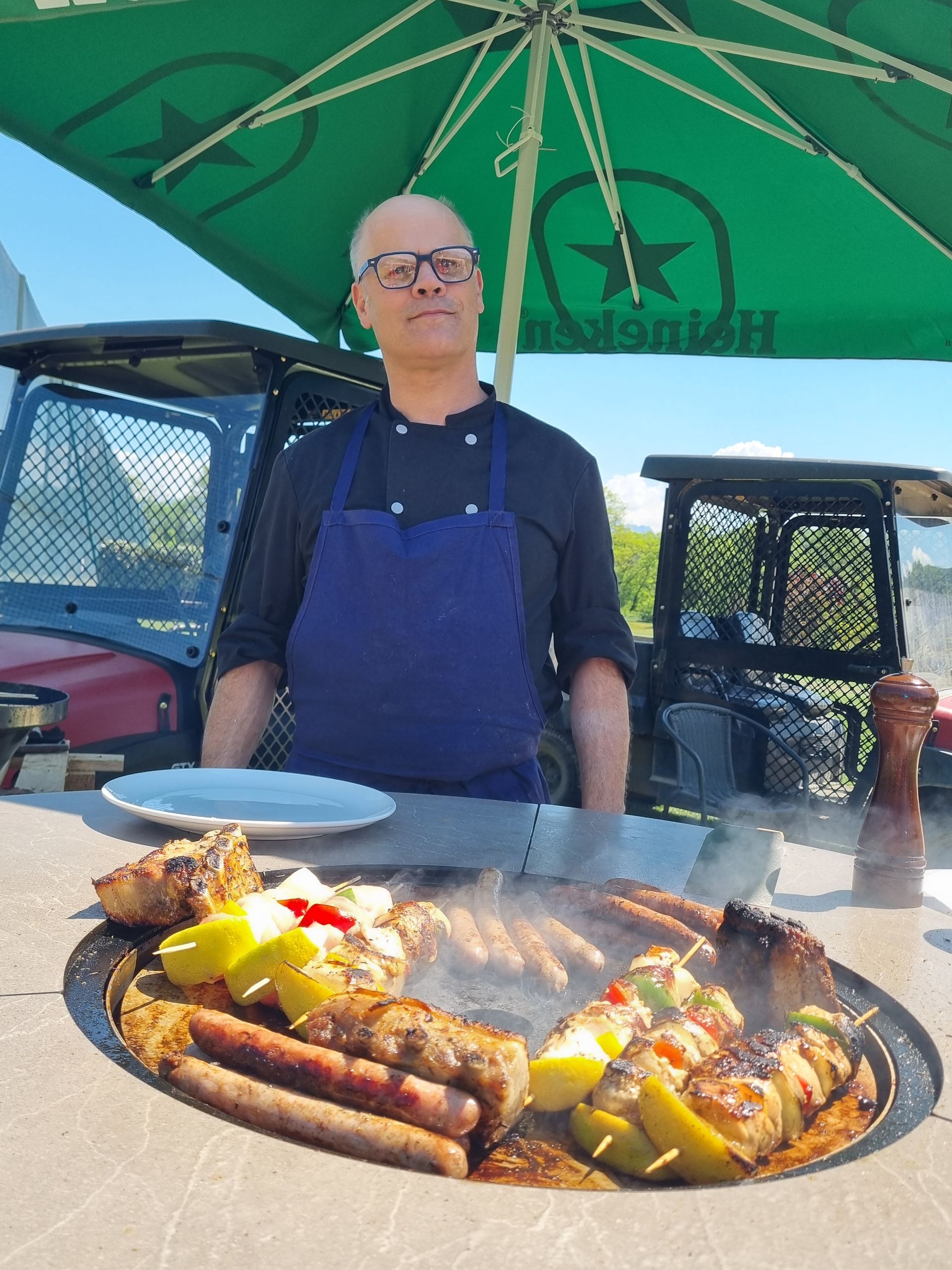 Une personne se tient derrière une table avec des grillades sous un parasol vert, à l'extérieur.