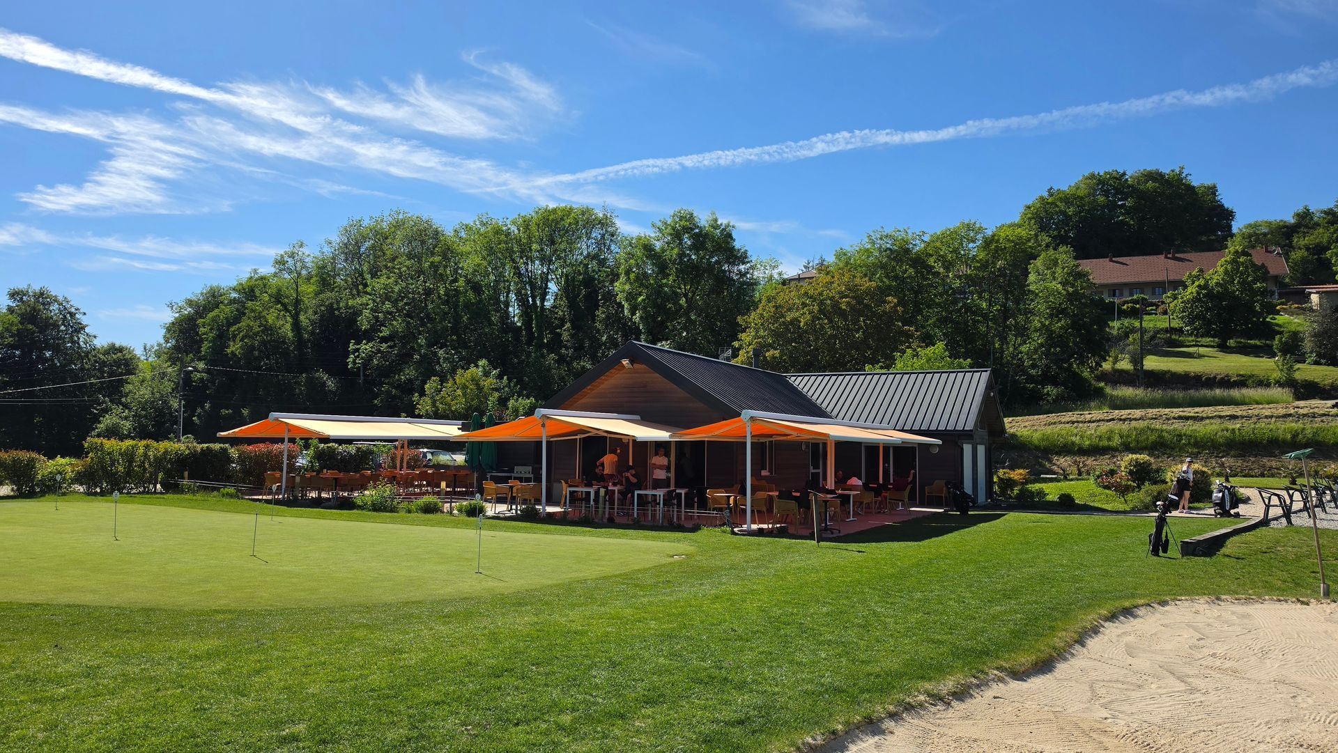 Café en plein air avec des parasols orange sur une pelouse verte ensoleillée, adossée à des arbres et à une maison à flanc de colline.
