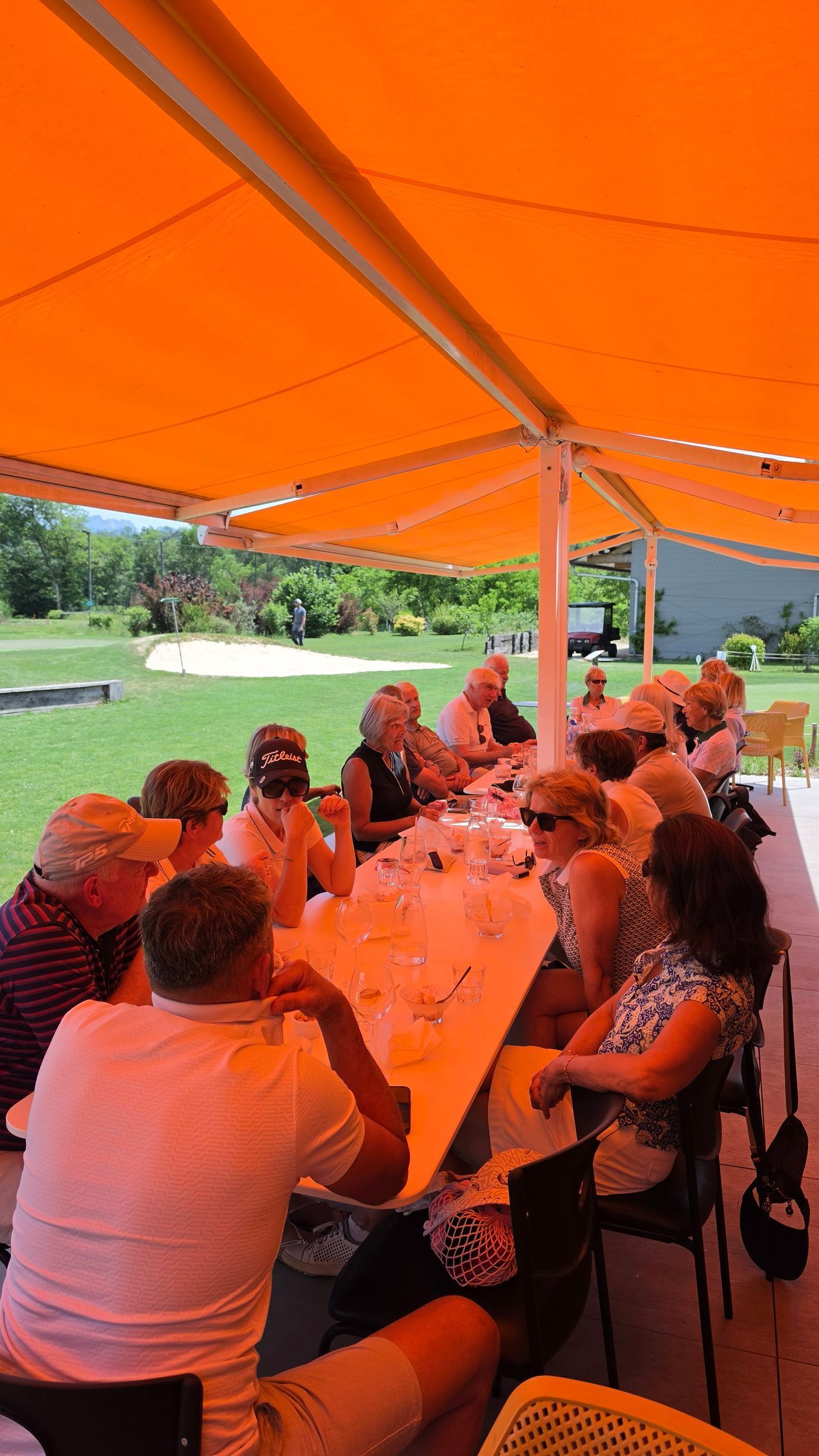 Des personnes assises à une longue table sous un auvent orange, mangeant et discutant en plein air.