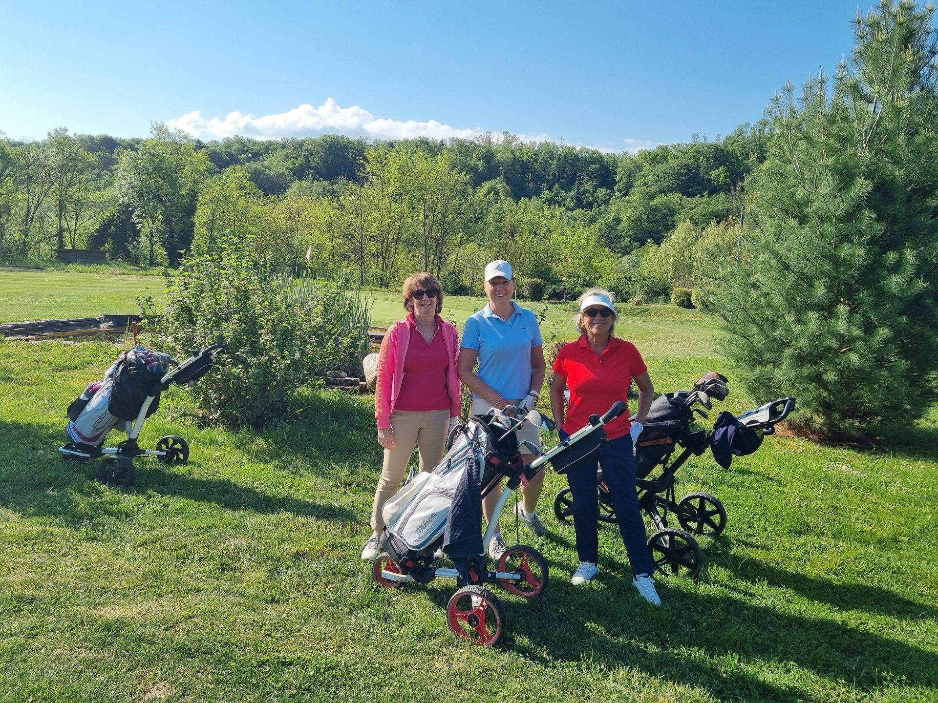Trois personnes posent avec des voiturettes de golf sur une pelouse verte ensoleillée, avec des arbres et des collines en arrière-plan.
