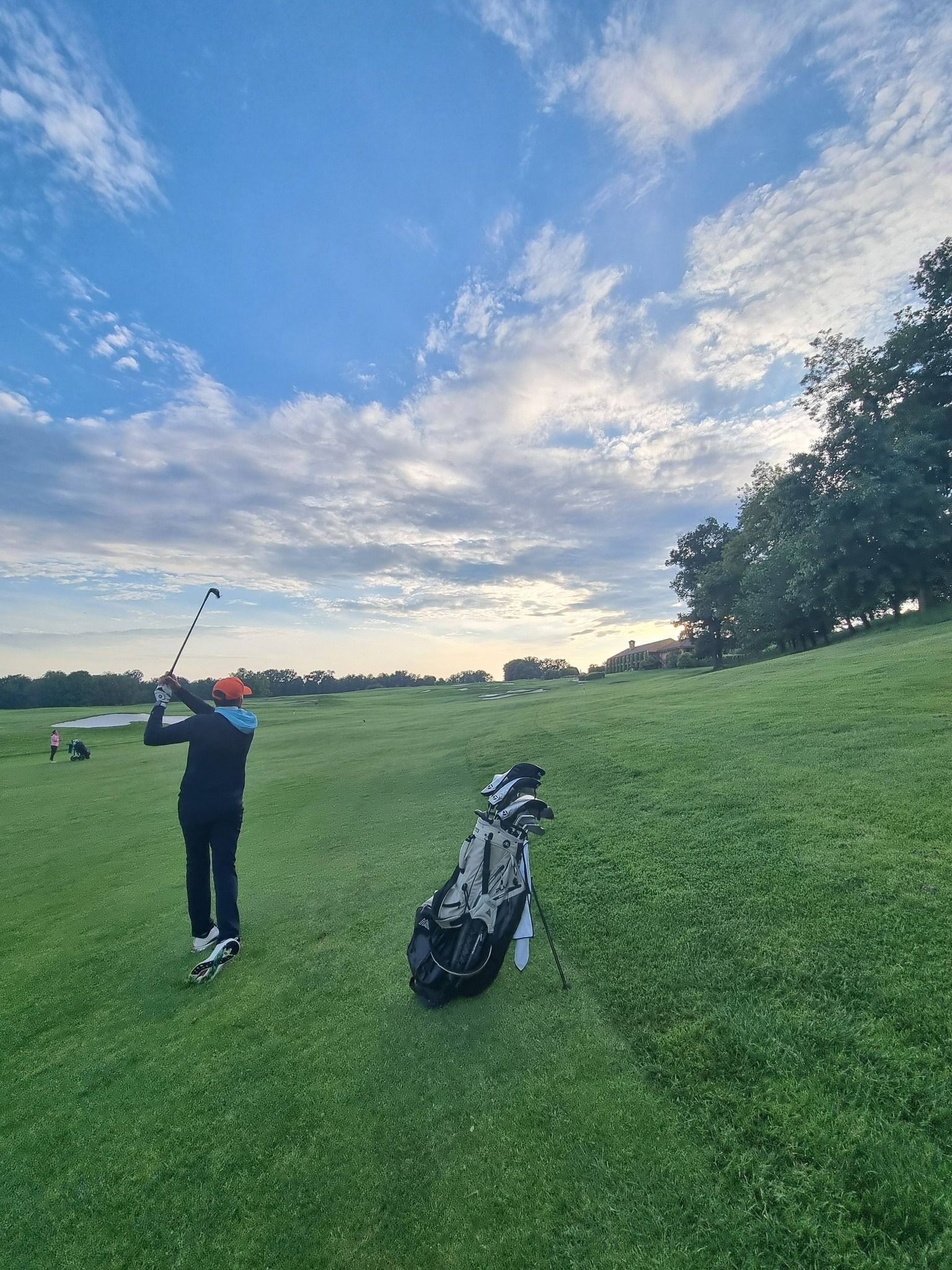 Un golfeur effectue un swing sur un parcours herbeux au coucher du soleil, à côté de son sac de golf, sous un ciel bleu nuageux.