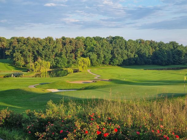Terrain de golf verdoyant et ensoleillé, avec un chemin sinueux, des arbres et des fleurs rouges au premier plan.