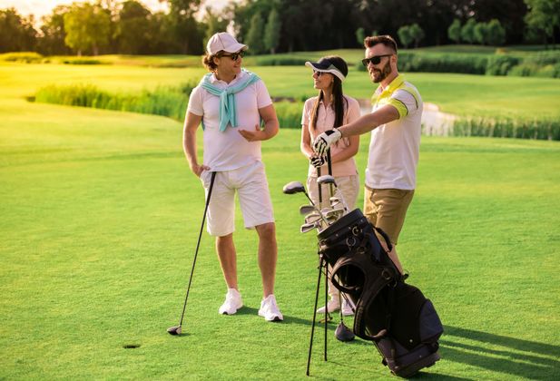 Trois golfeurs se tiennent debout sur un fairway verdoyant et ensoleillé, clubs et sac de golf à la main.