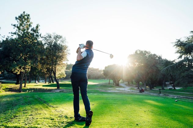 Un golfeur effectue un swing sur un parcours verdoyant au coucher du soleil, avec des arbres et un soleil éclatant en arrière-plan.