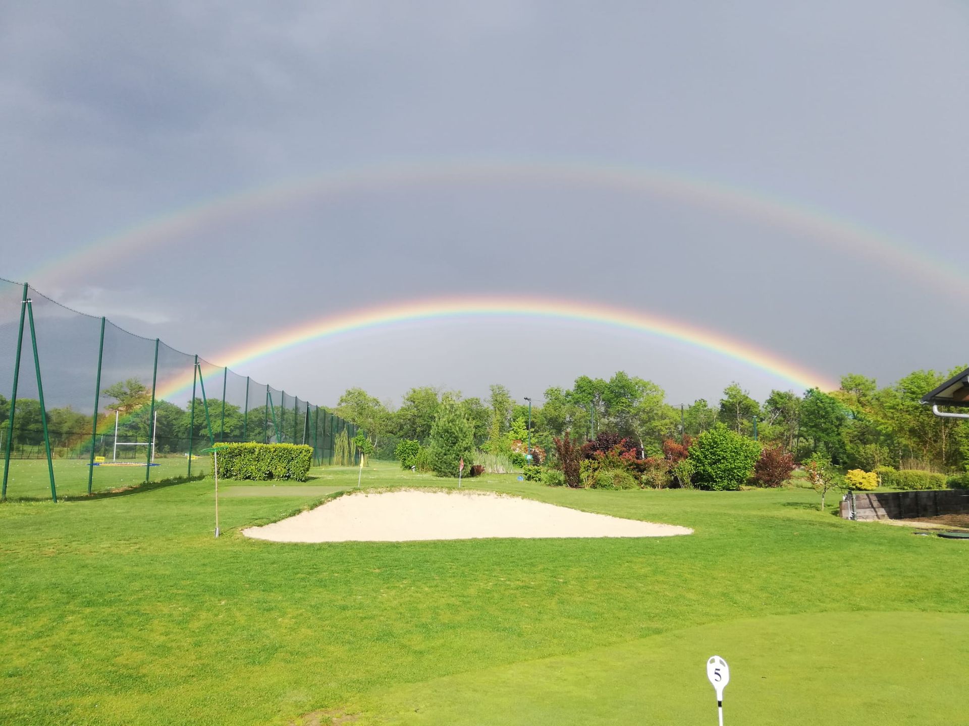 Terrain de golf avec un green sous un double arc-en-ciel, un bunker de sable et des arbres sous un ciel nuageux.